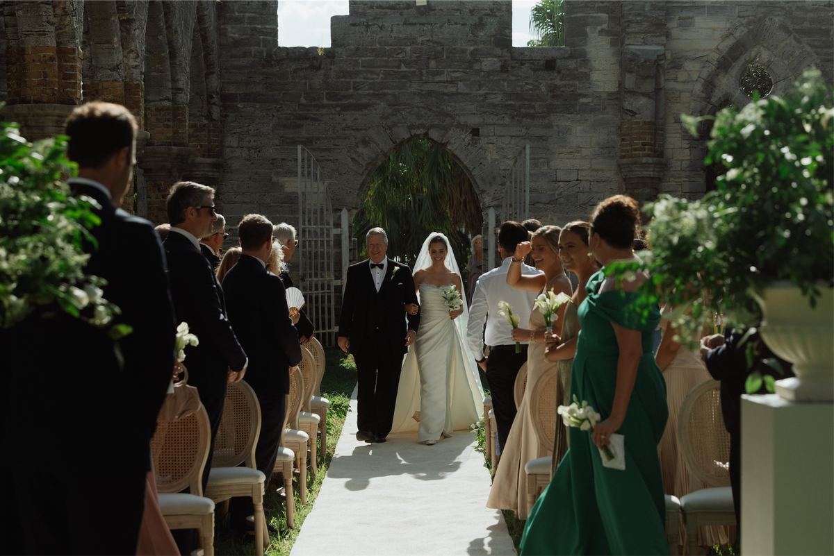 Bride and her father walking the aisle toward the stone arch as guests stand