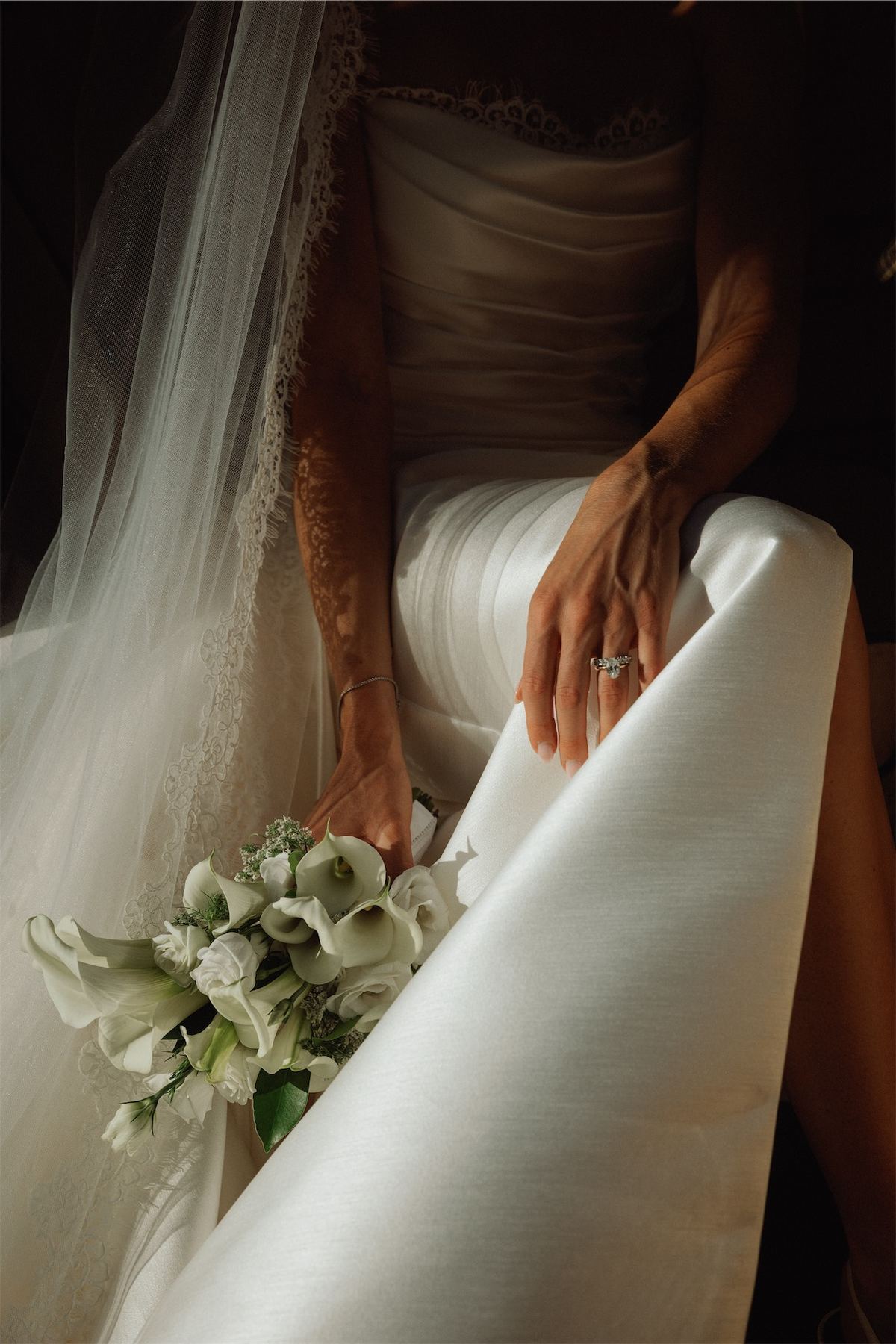 Detail of the bride's hands gathering her satin gown beside a calla lily bouquet