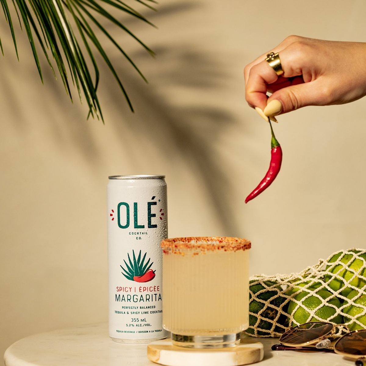 Hand dropping a red chilli into a salt-rimmed spicy margarita beside its can