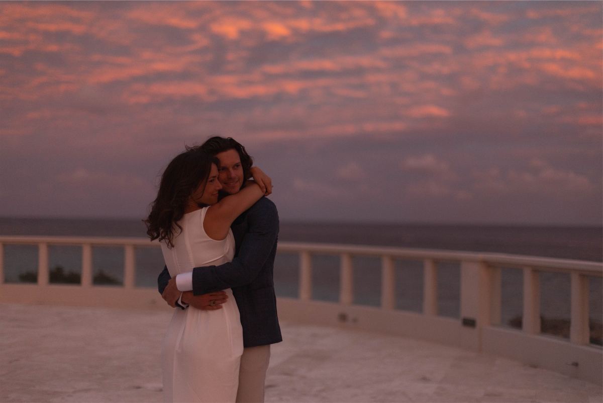 Couple holding each other against a pink and orange sunset over the water