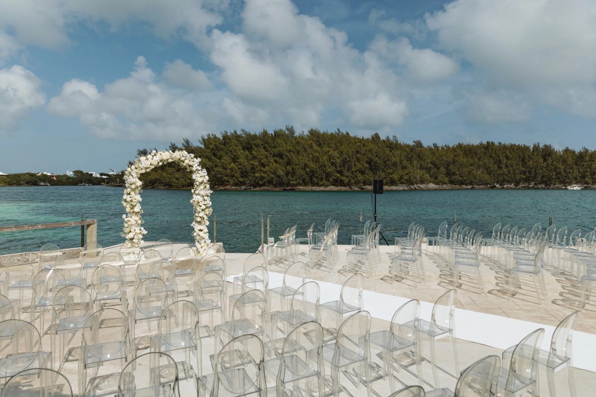 Clear ceremony chairs facing a white floral arch over the water on Hawkins Island, Bermuda