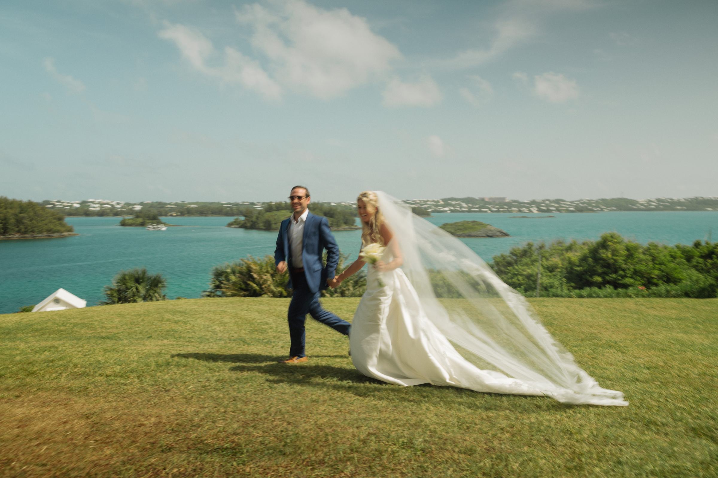 Paige and Cam walk the clifftop lawn above the bay, her veil streaming in the wind on Hawkins Island, Bermuda