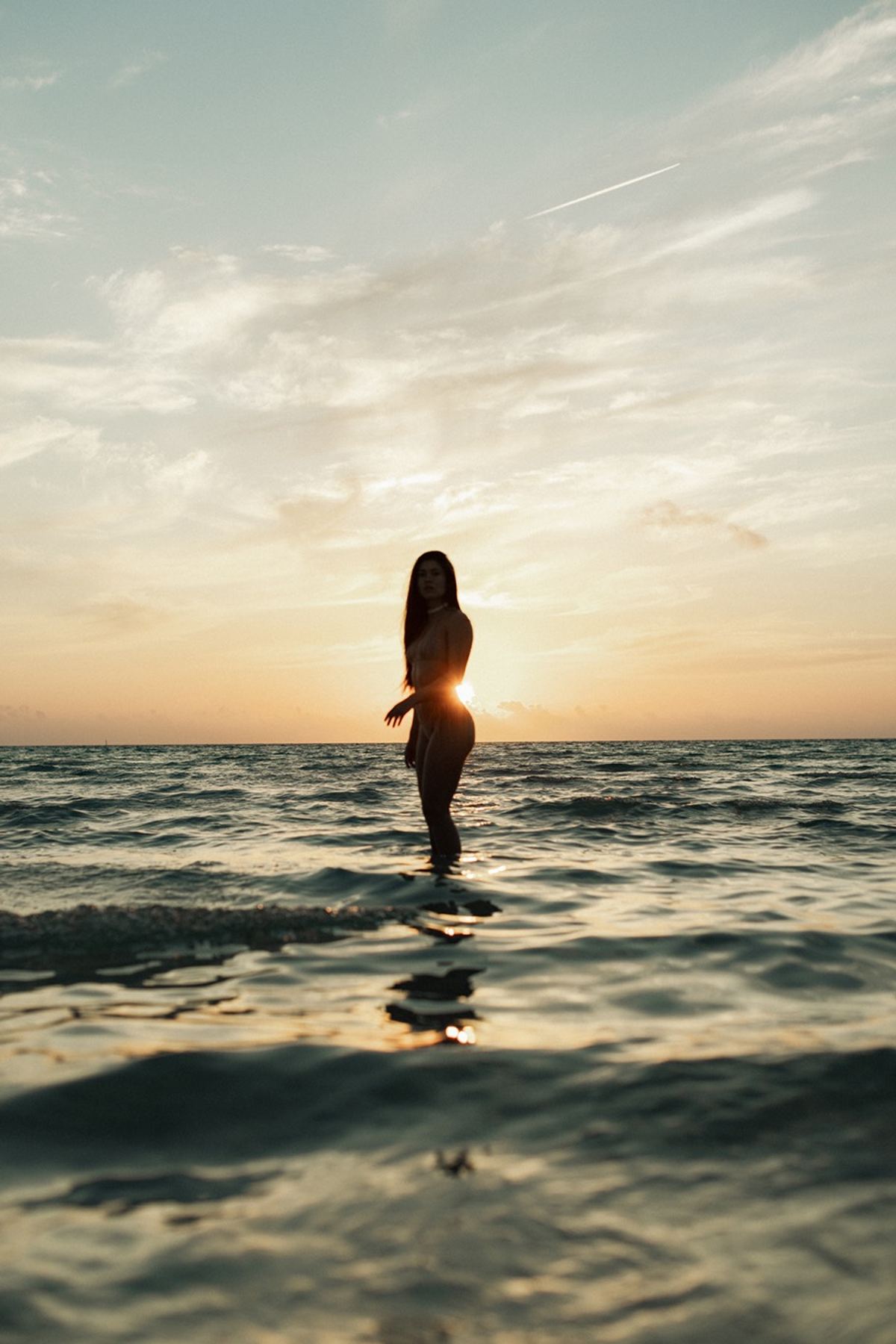 Swimwear model standing waist deep in the ocean, backlit by a low sunset