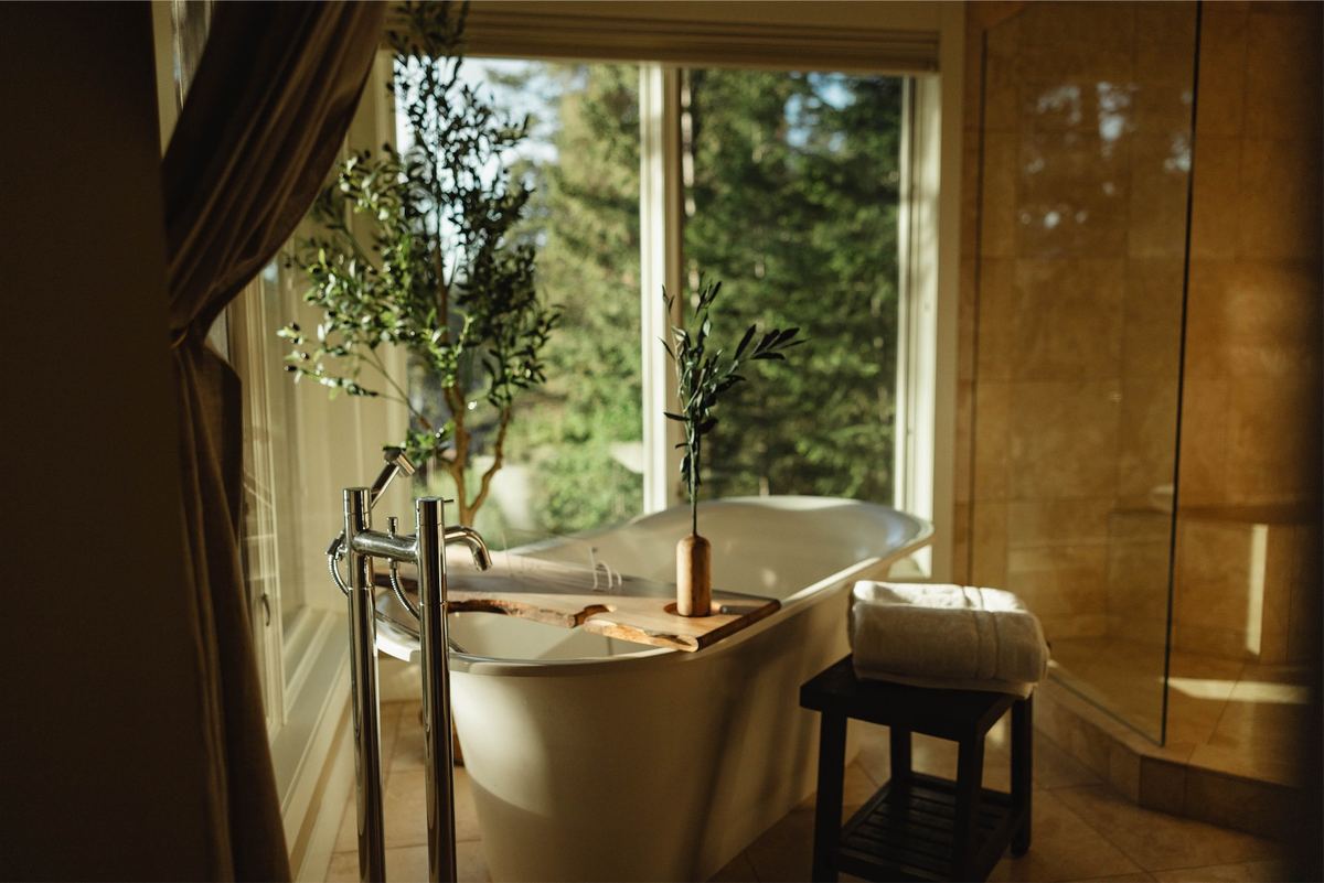 Freestanding tub beside a window framed by olive trees and forest