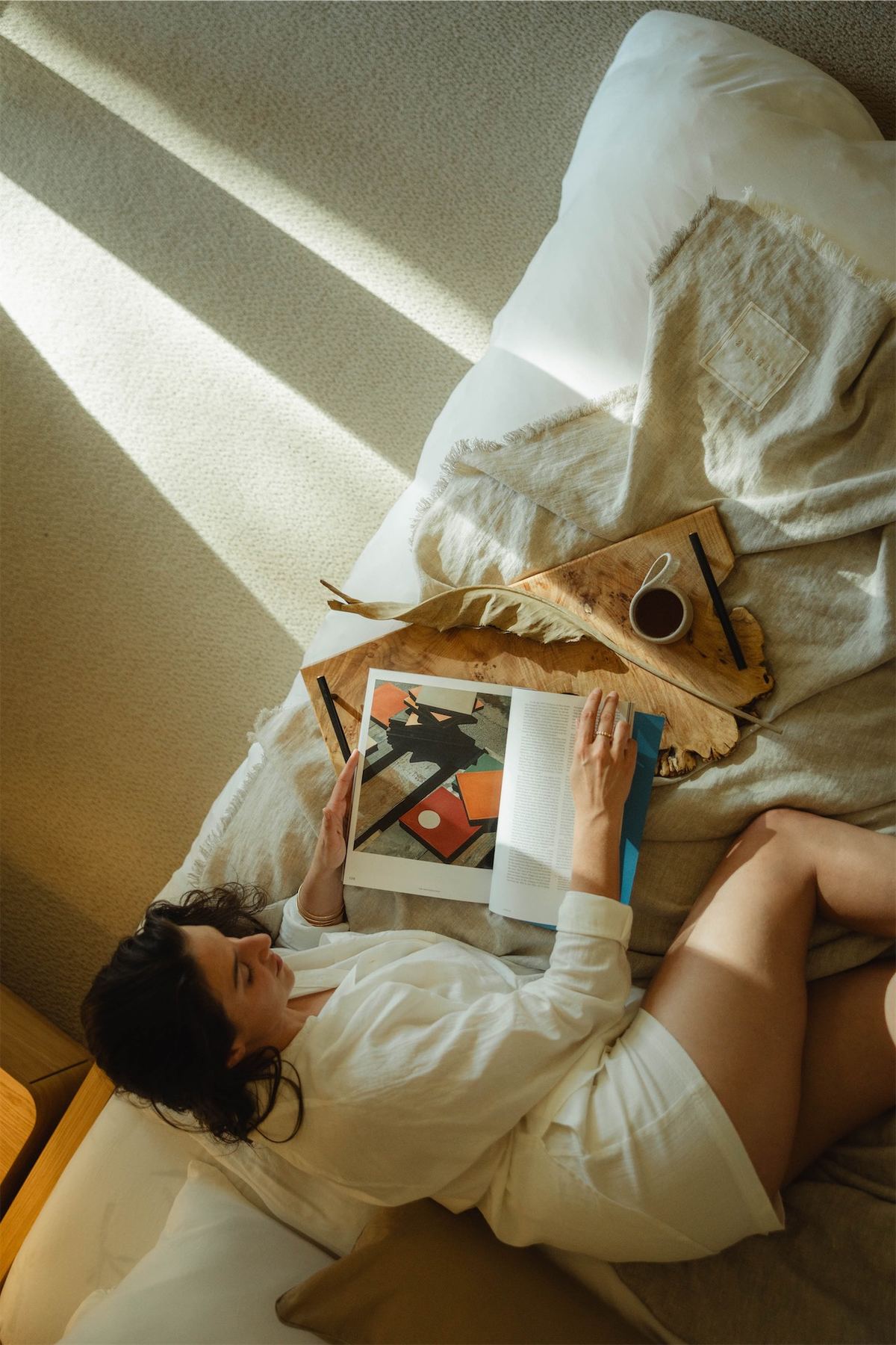 Overhead of a woman in a white robe reading in bed with coffee and a book