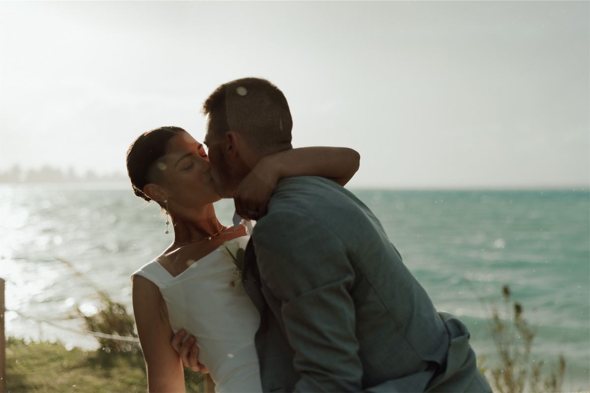 Bride and groom kissing on a grassy point with open ocean behind them