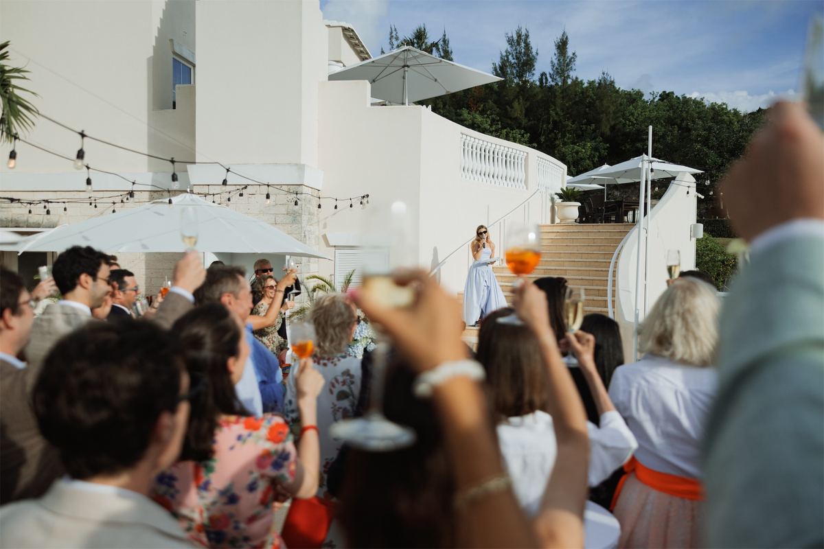 Guests lifting cocktails and cheering as the couple appear above a poolside terrace