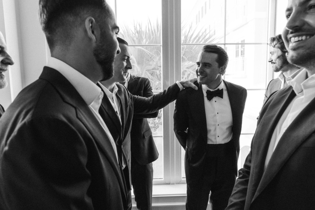 Groom laughing with his groomsmen by the window at Rosewood Bermuda, black and white