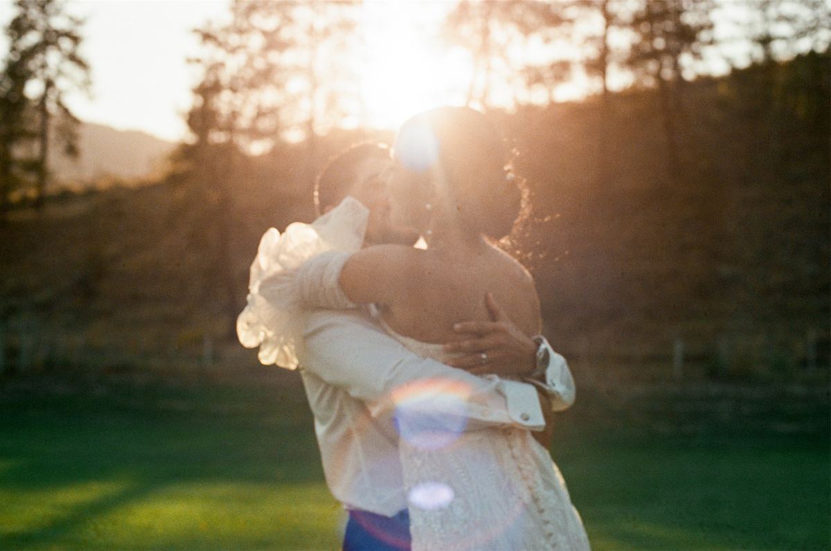 Couple embracing in an open field as the sun flares behind the treeline
