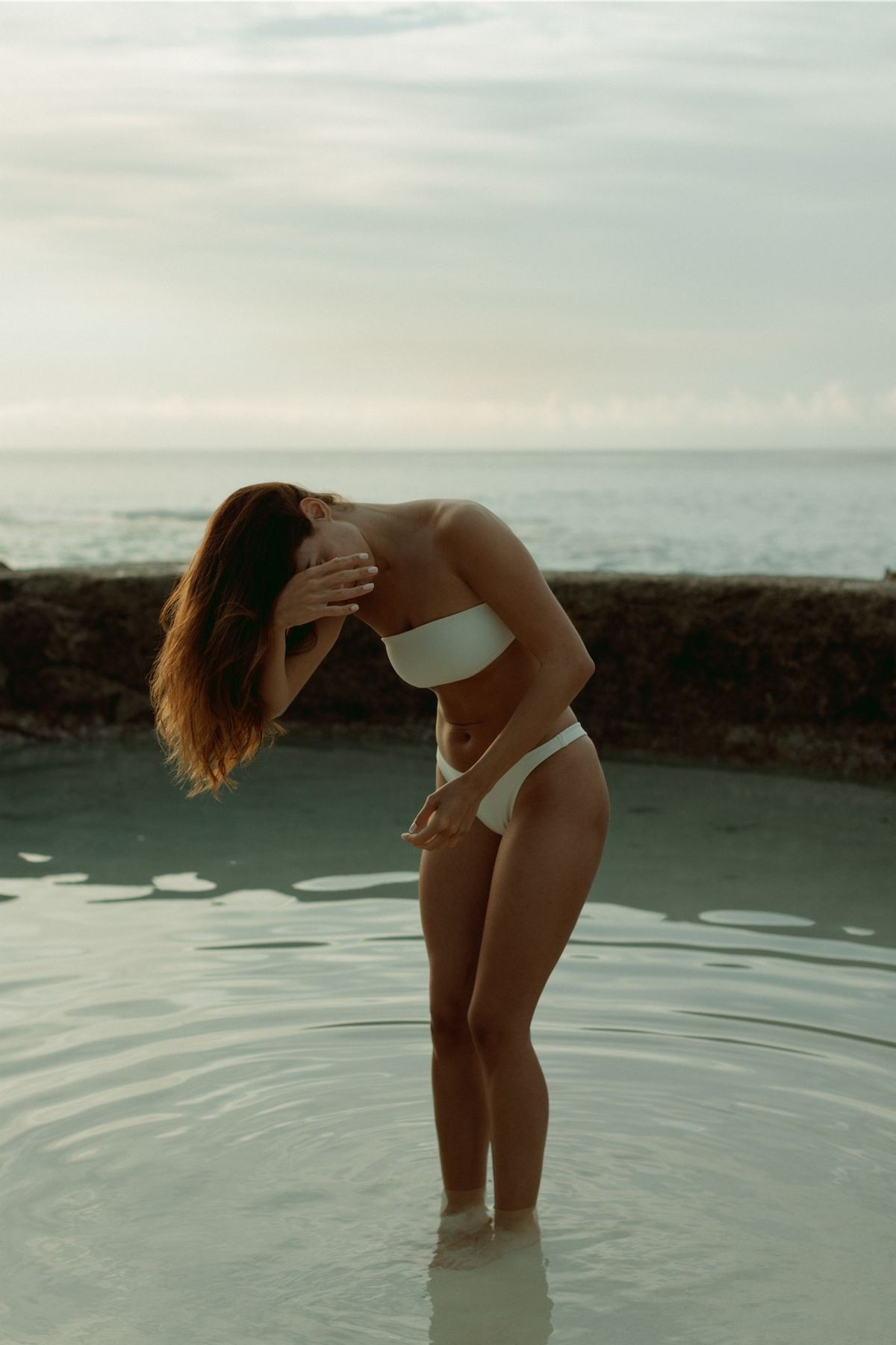 Model in a white bikini standing in a shallow tidal pool, hands lifted to her hair