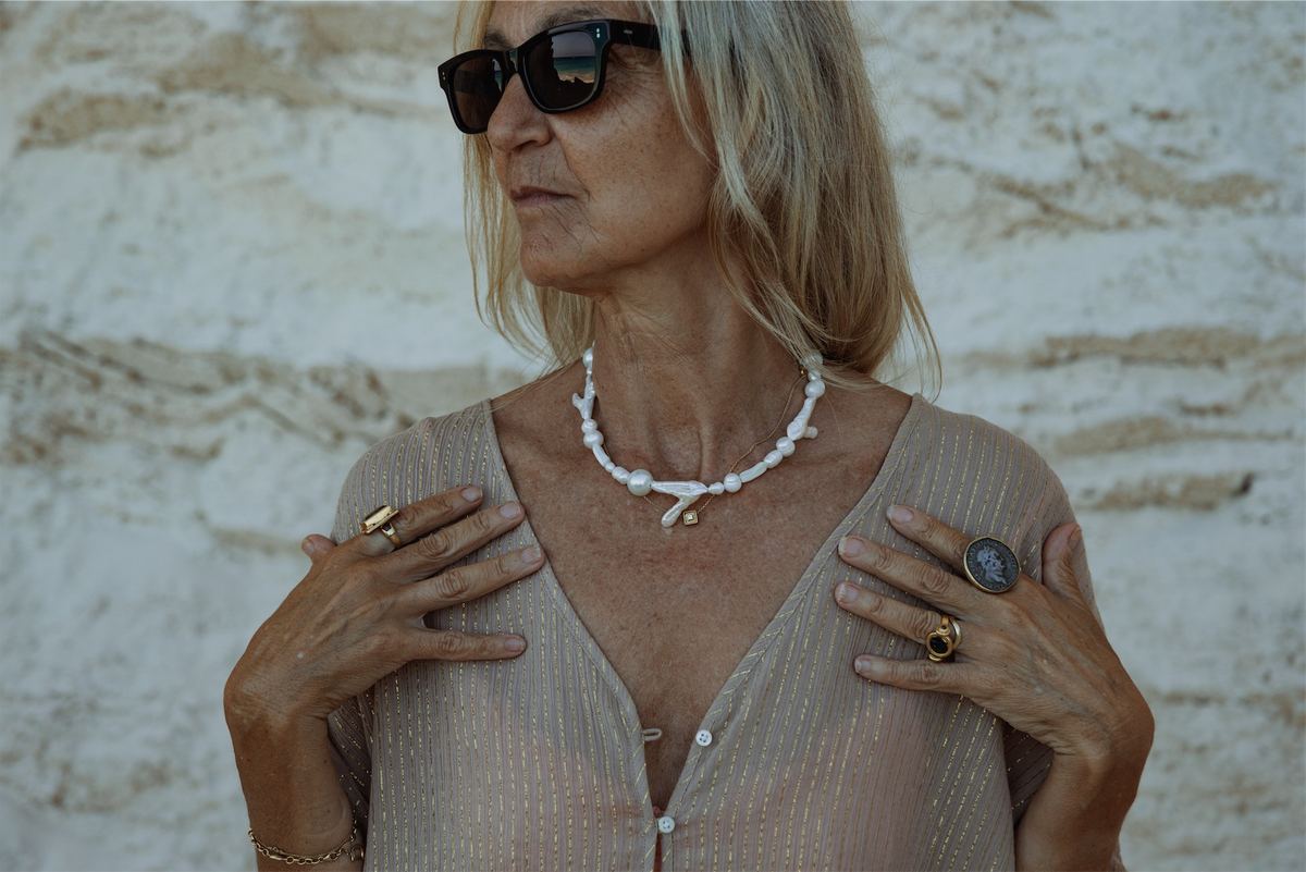 Portrait of a silver haired model in a shell necklace and gold rings against pale rock