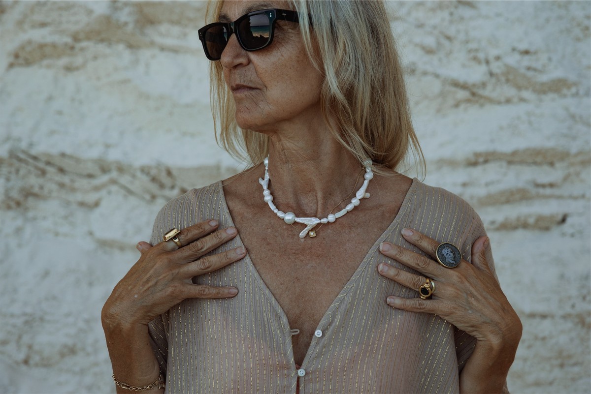 Portrait of a silver haired model in a shell necklace and gold rings against pale rock