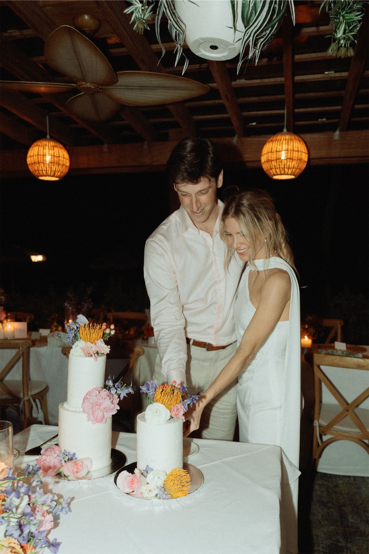 Couple cutting their tiered wedding cake under glowing lanterns at the reception