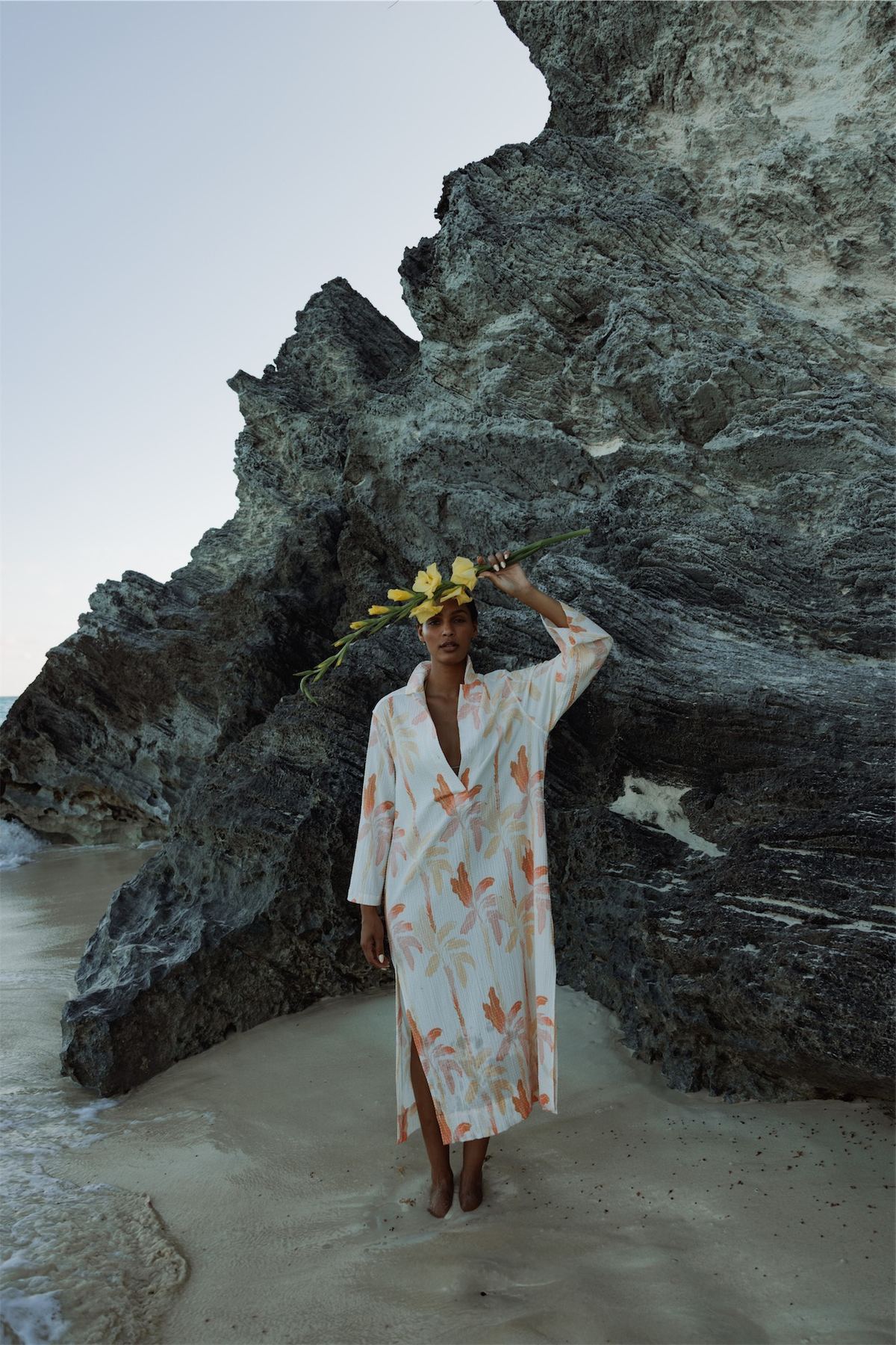 Model in a printed kaftan standing on the sand beneath a dark cliff face