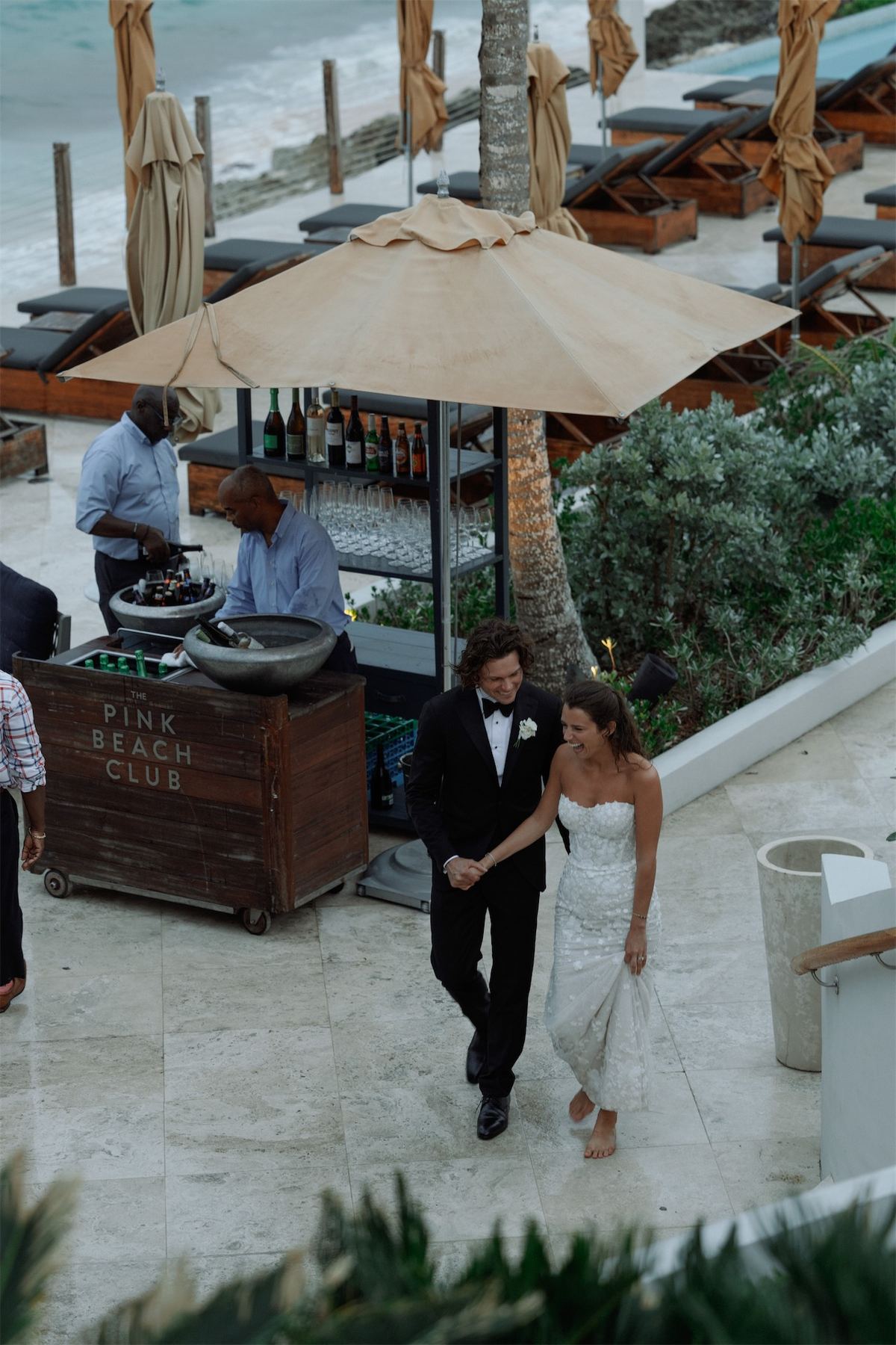Bride and groom walking past a beach club bar cart shaded by canvas umbrellas