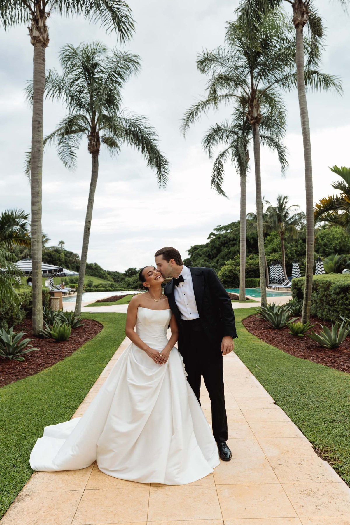 Groom kissing the bride's temple on the garden path under tall palms at Rosewood Bermuda