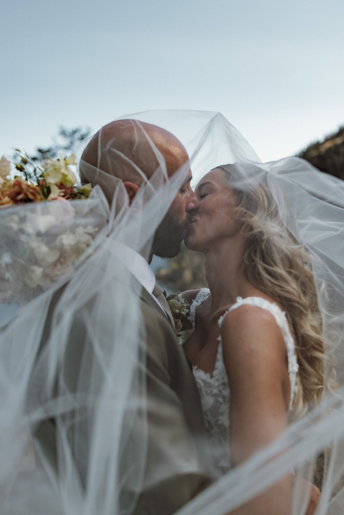Bride and groom kissing beneath a wind blown veil above the lake