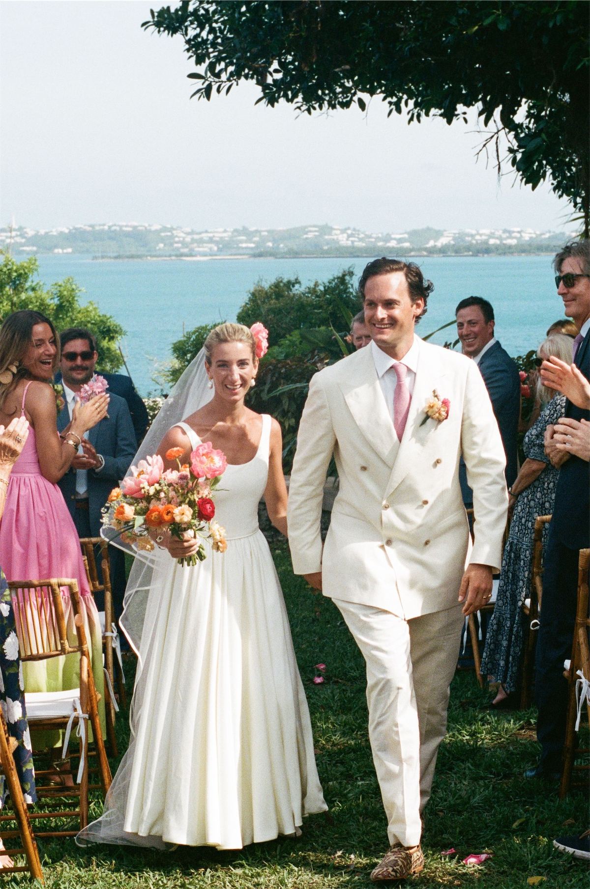 Newlyweds walking back down the aisle as guests applaud, ocean behind them