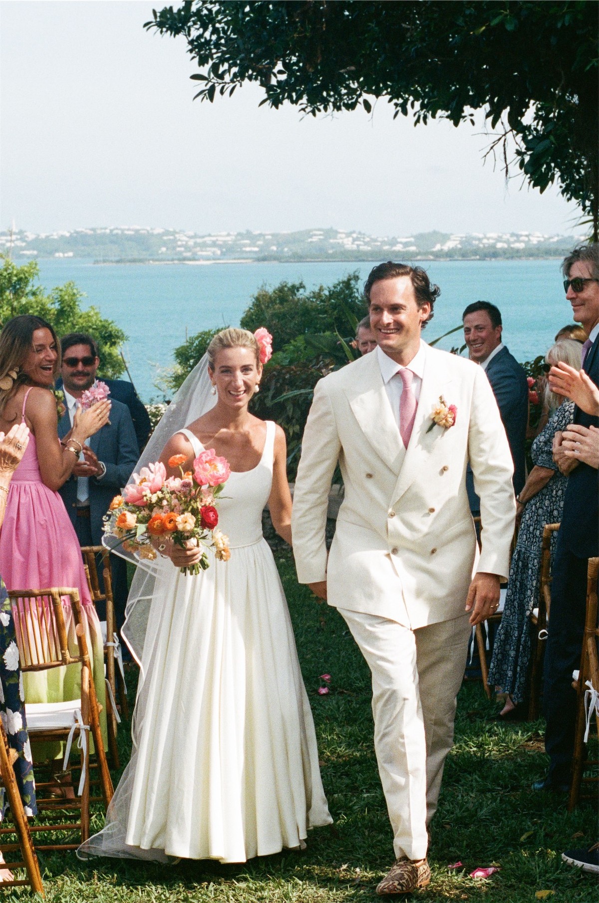 Newlyweds walking back down the aisle as guests applaud, ocean behind them