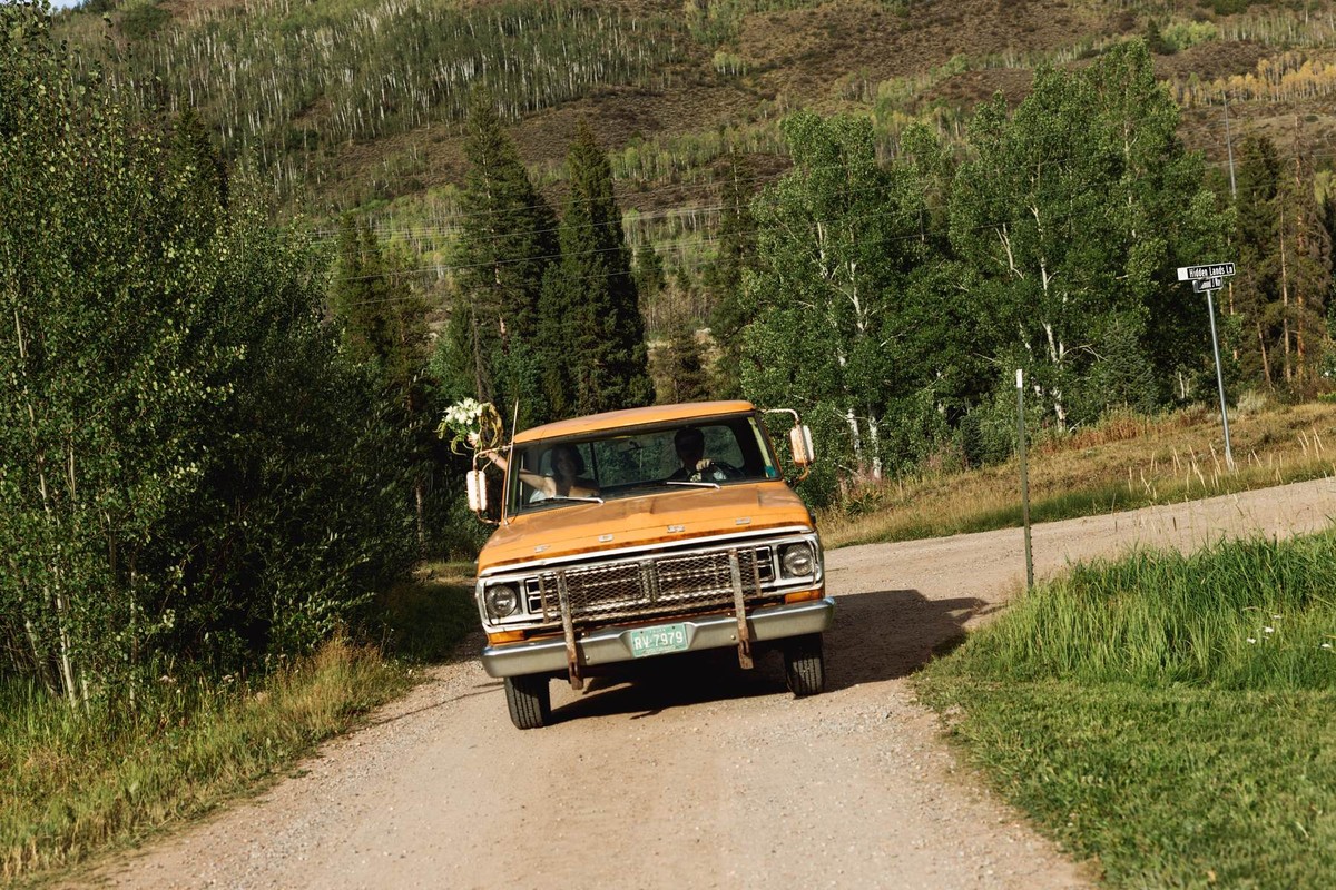 The orange pickup drives down the dirt road with a bouquet on the mirror at a mountain ranch among the aspens