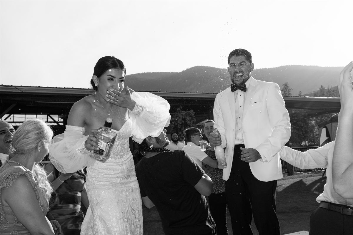 Black and white of the bride swigging from a bottle as guests celebrate around her