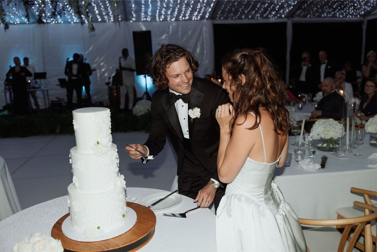 Bride and groom cutting a tiered white wedding cake inside the tented reception