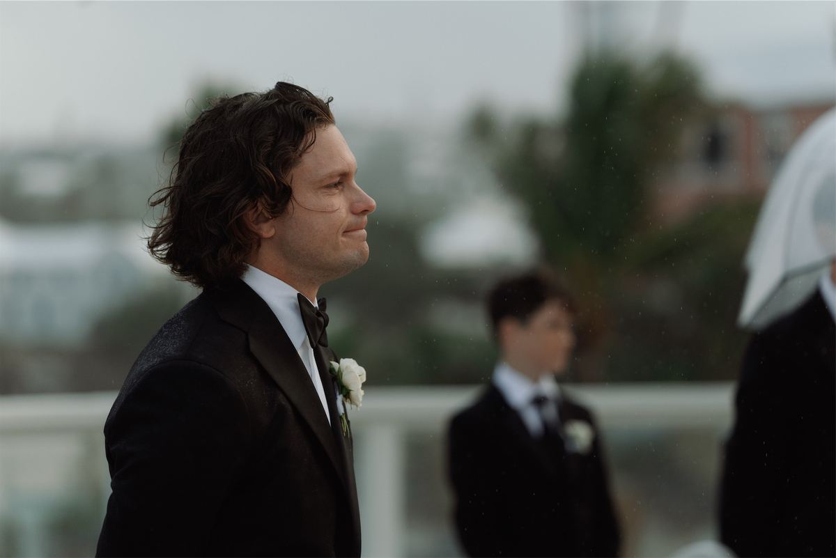 Groom in a black tuxedo watching the ceremony, groomsmen blurred behind him