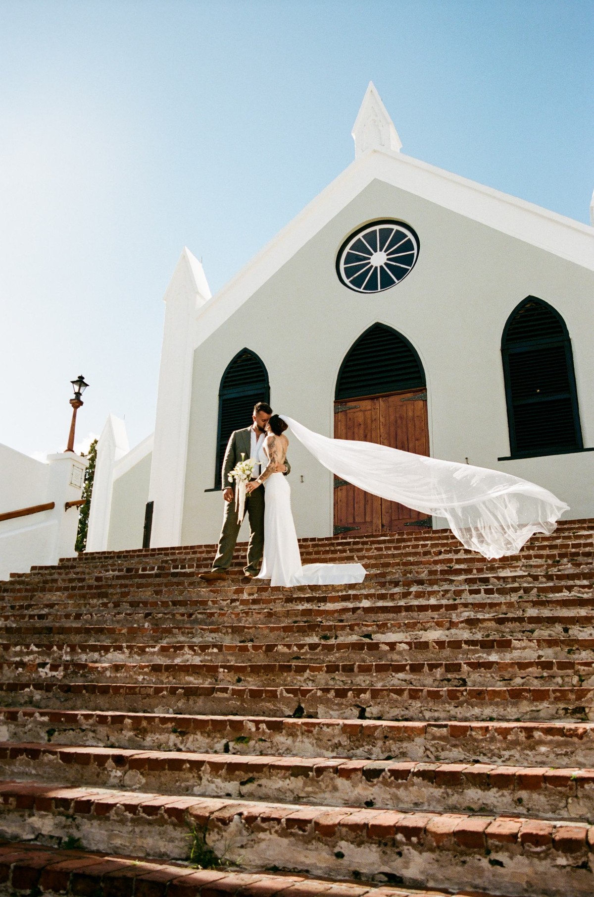 Bride and groom on the church steps with her veil caught by the wind, shot on film