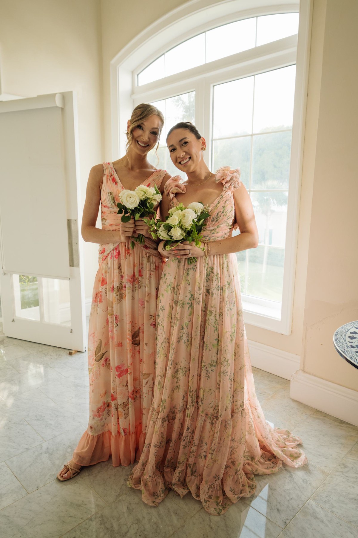 Two bridesmaids in floral gowns smiling by an arched window at Rosewood Bermuda