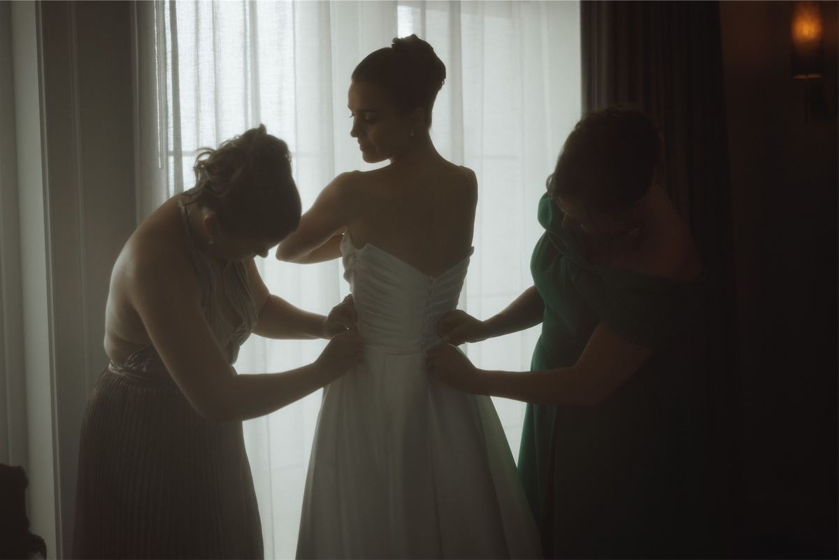 Bridesmaids buttoning the bride into her gown beside a bright window