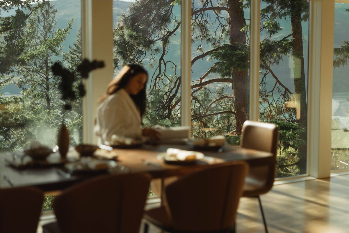Woman in a robe at the dining table with the pines through the window