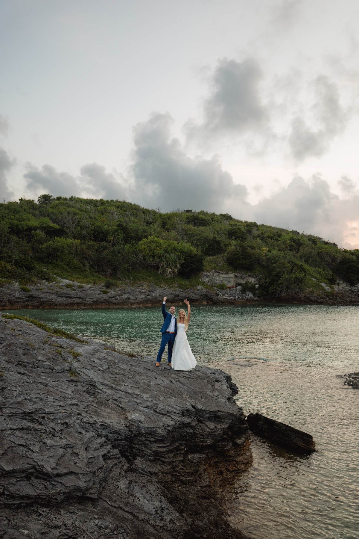 Paige and Cam stand on the rocks at the water's edge at dusk on Hawkins Island, Bermuda