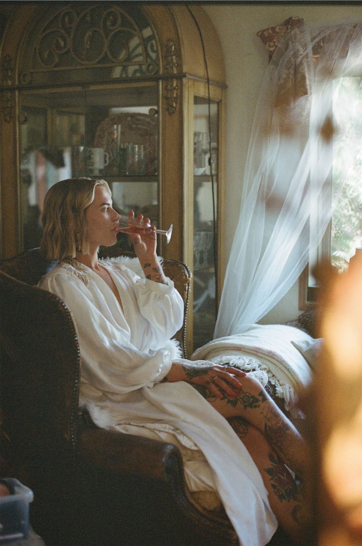 Bride sipping a drink beside a bright window while getting ready