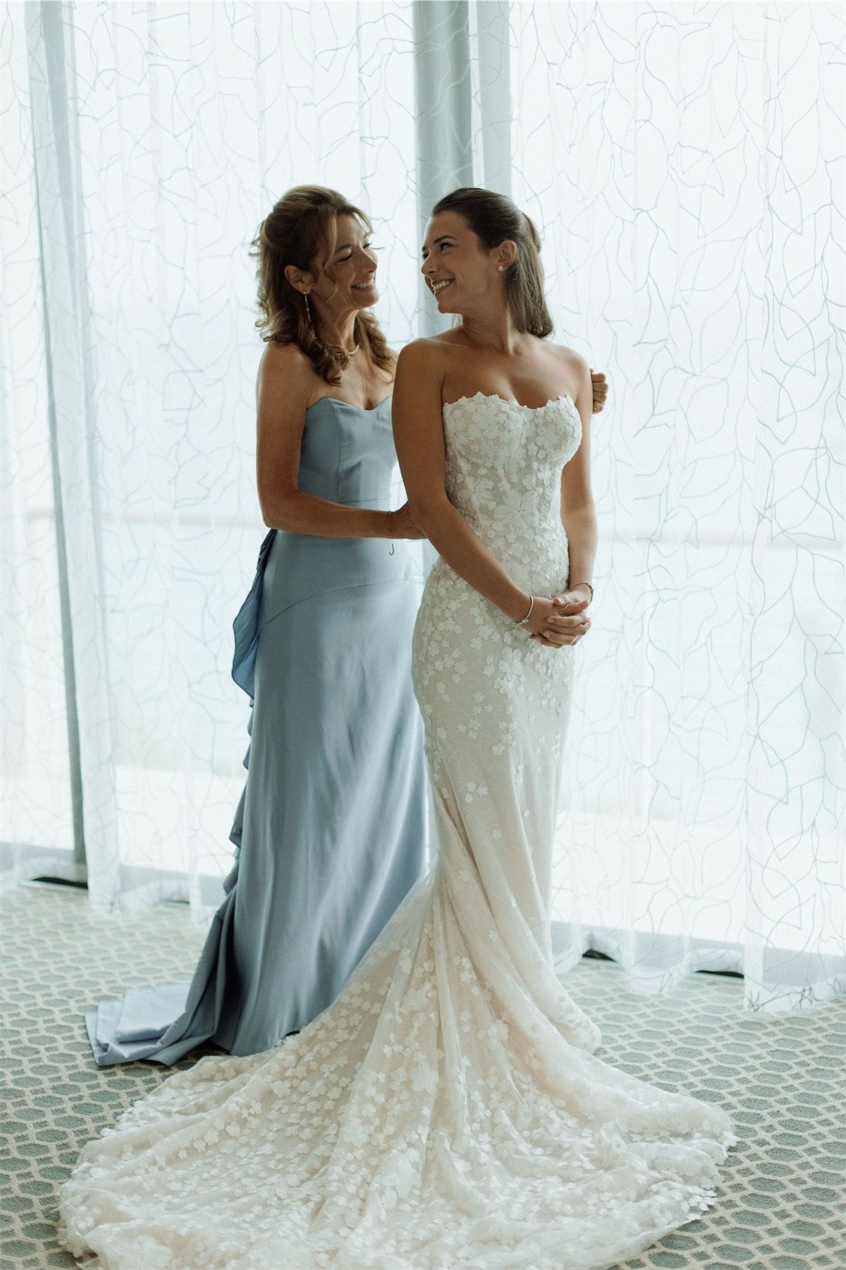 Bride and her mother in blue standing together beside a sheer curtained window