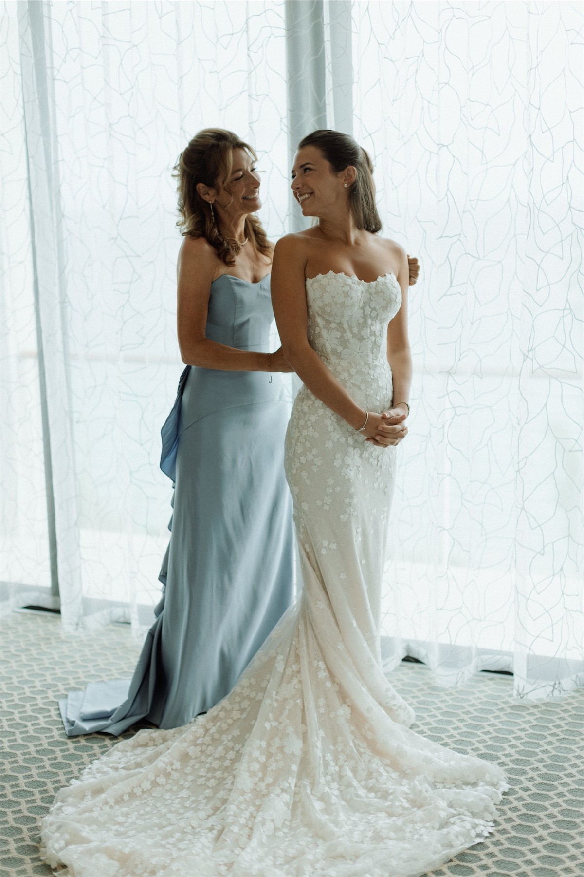 Bride and her mother in blue standing together beside a sheer curtained window