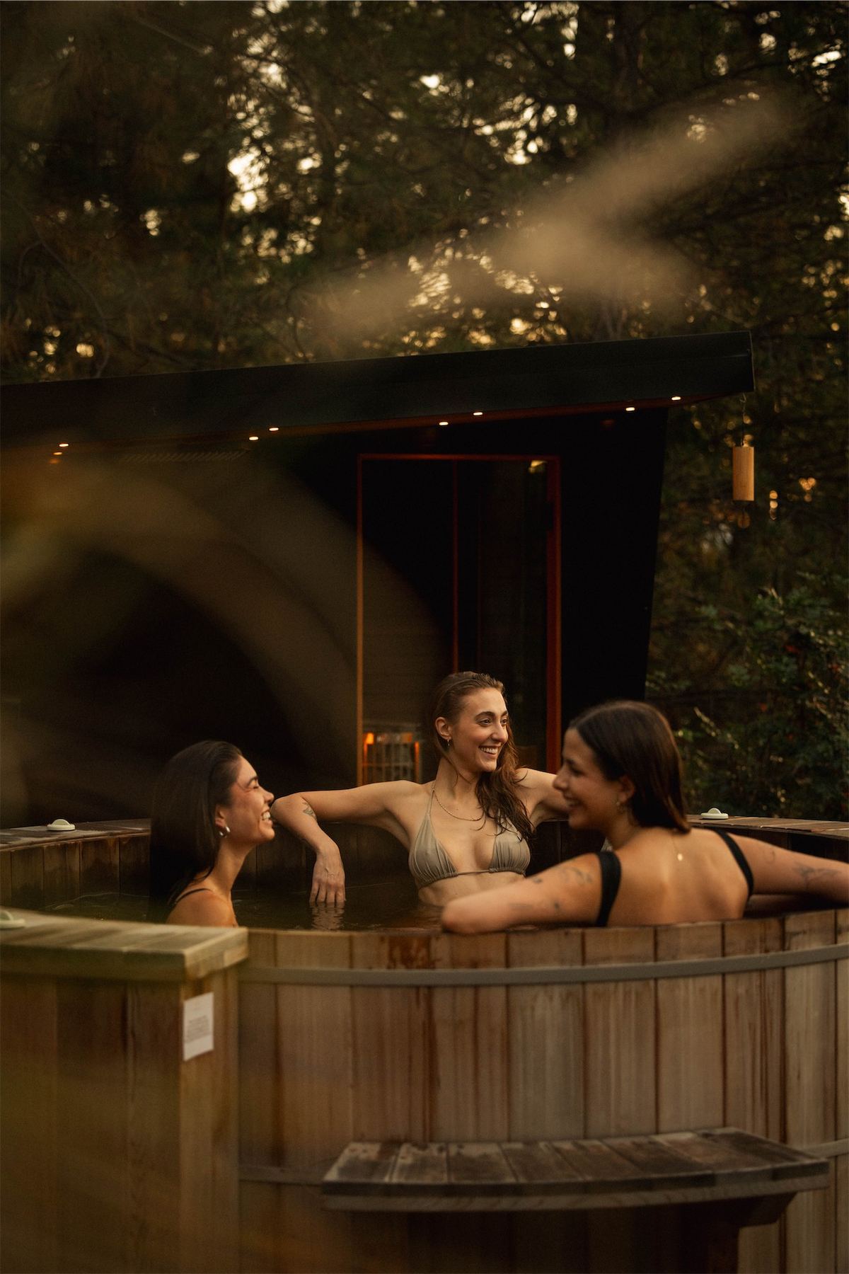 Three friends laughing together in a cedar hot tub outside the sauna
