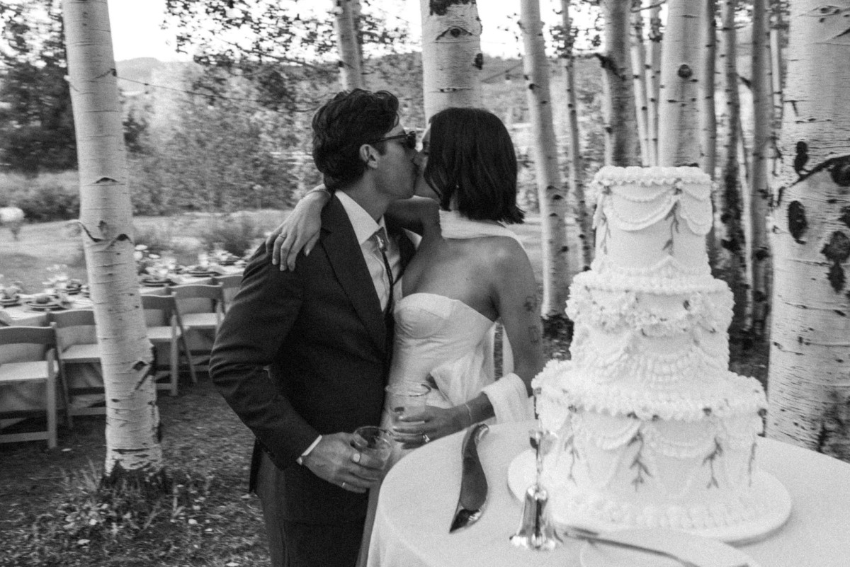 Owen kisses Devin beside their wedding cake in the aspens at a mountain ranch among the aspens, black and white