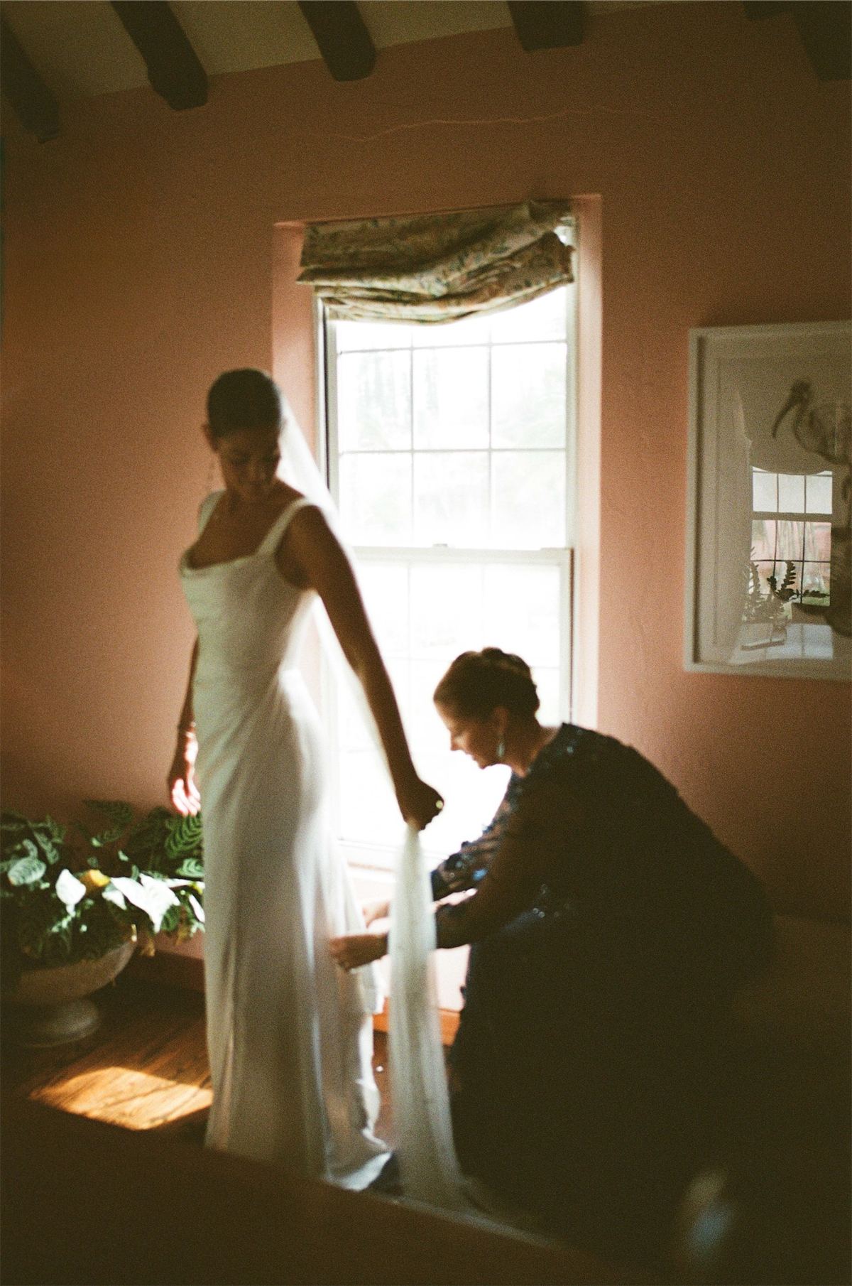 Bride having her gown adjusted at a sunlit window in a pink bedroom