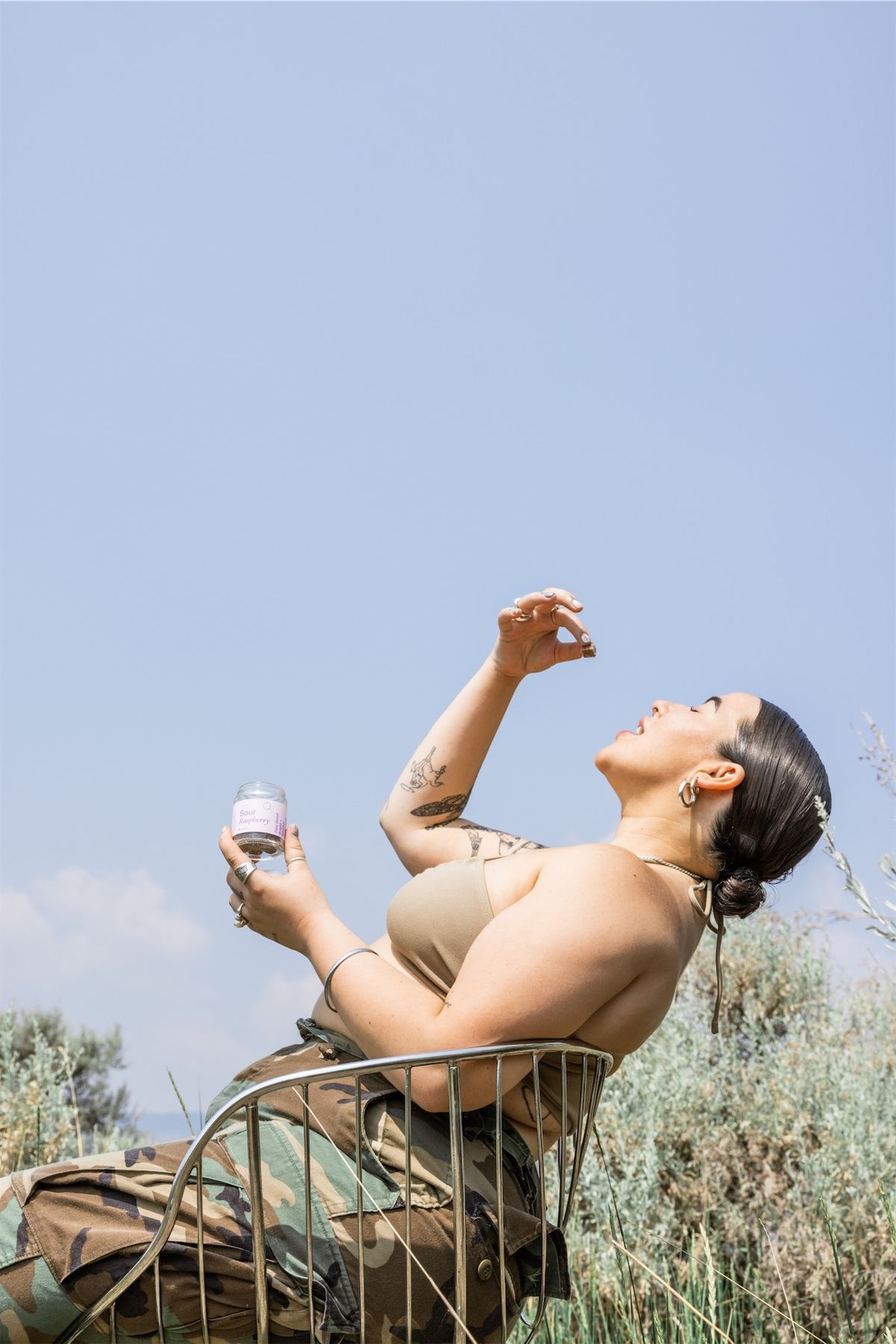 Model reclining in a wire chair in desert scrub, lifting a capsule jar to the sky