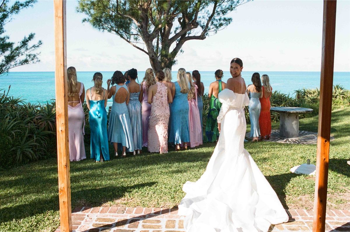 Bride turns to reveal her gown to the bridesmaids on a lawn above the ocean