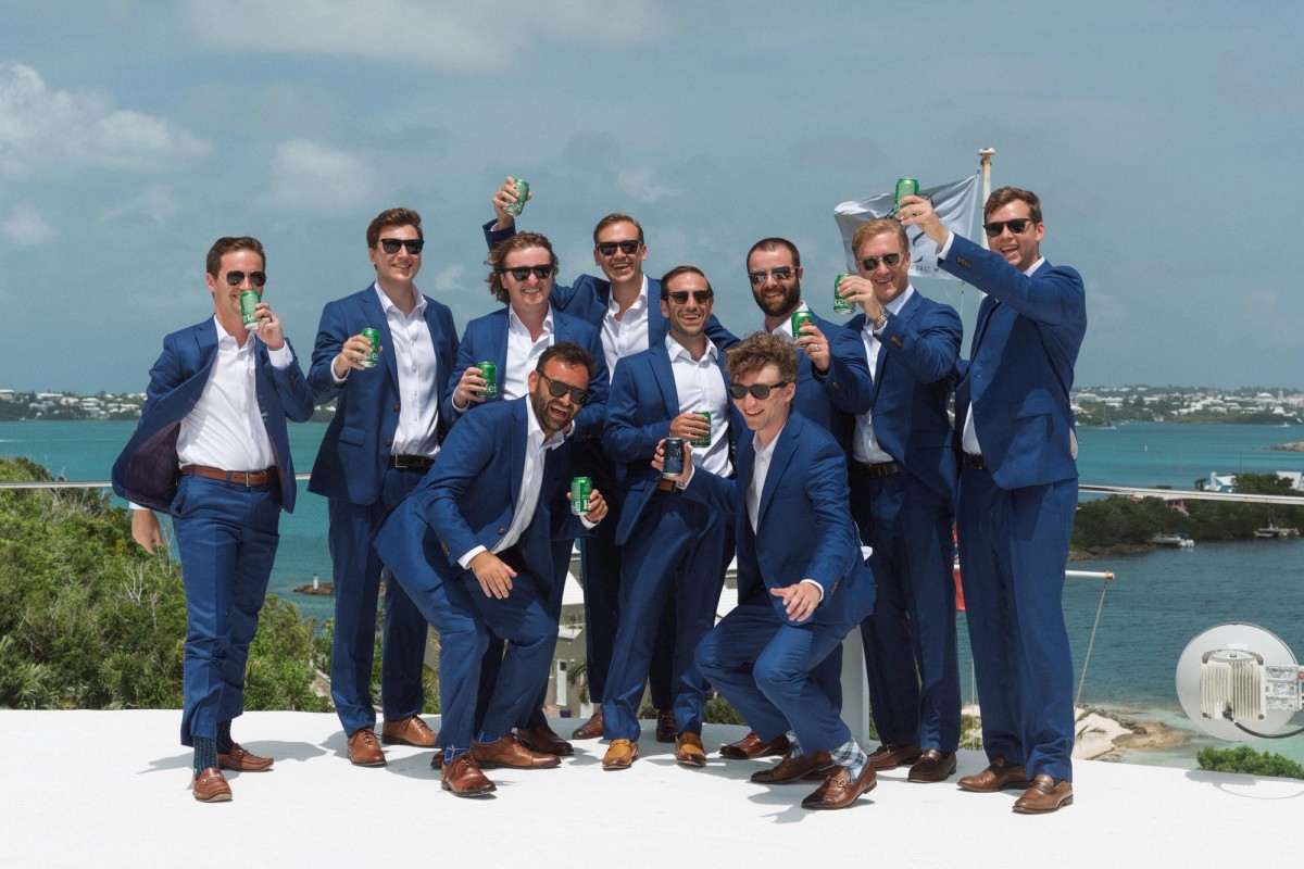 Groomsmen in blue suits raise drinks on the rooftop above the harbour on Hawkins Island, Bermuda