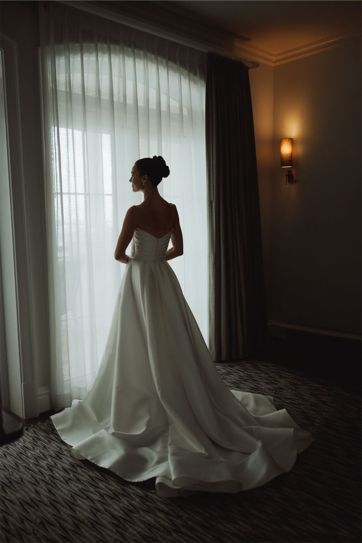 Bride in a full satin gown standing in window light with her train fanned out