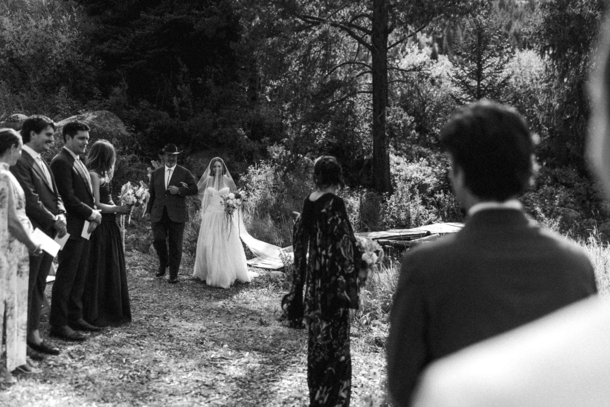 Devin walks down the aisle with her father as Owen waits at a mountain ranch among the aspens, black and white