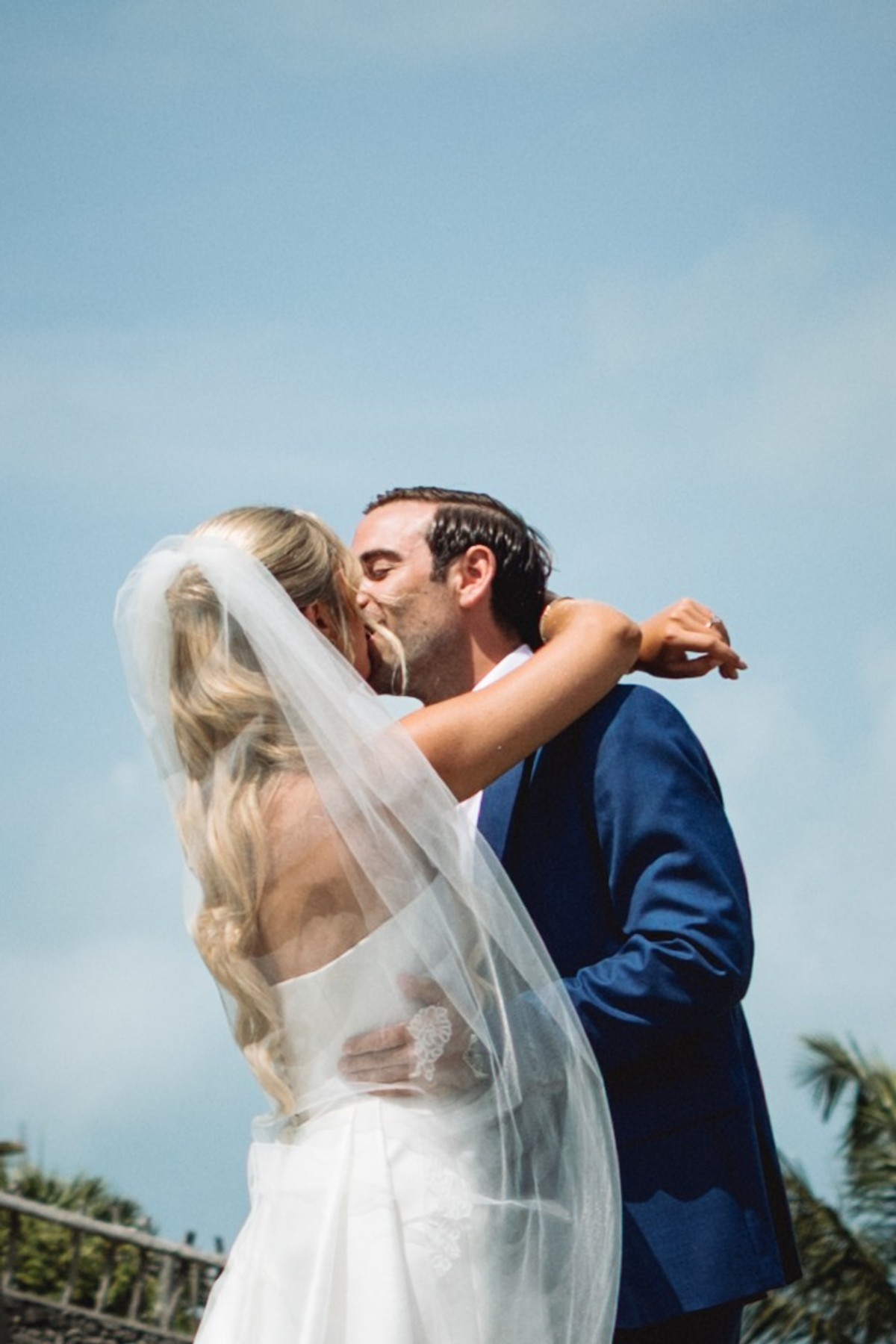 Cam kisses Paige under a clear blue sky, her veil wrapped around them on Hawkins Island, Bermuda