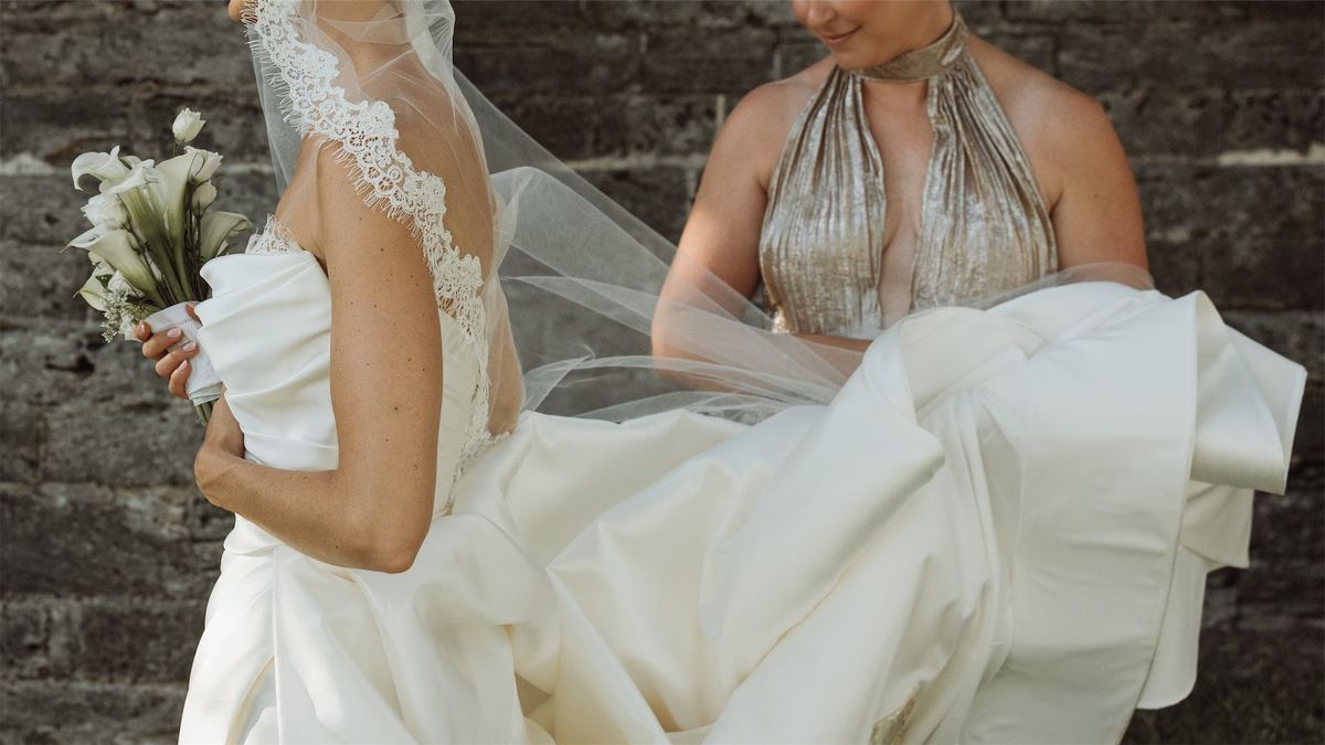 Bride in her veil embracing a guest in gold beside the stone wall