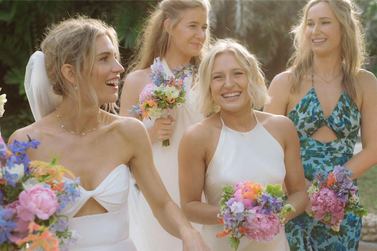 Bride laughing with bridesmaids in mismatched dresses holding bright spring bouquets