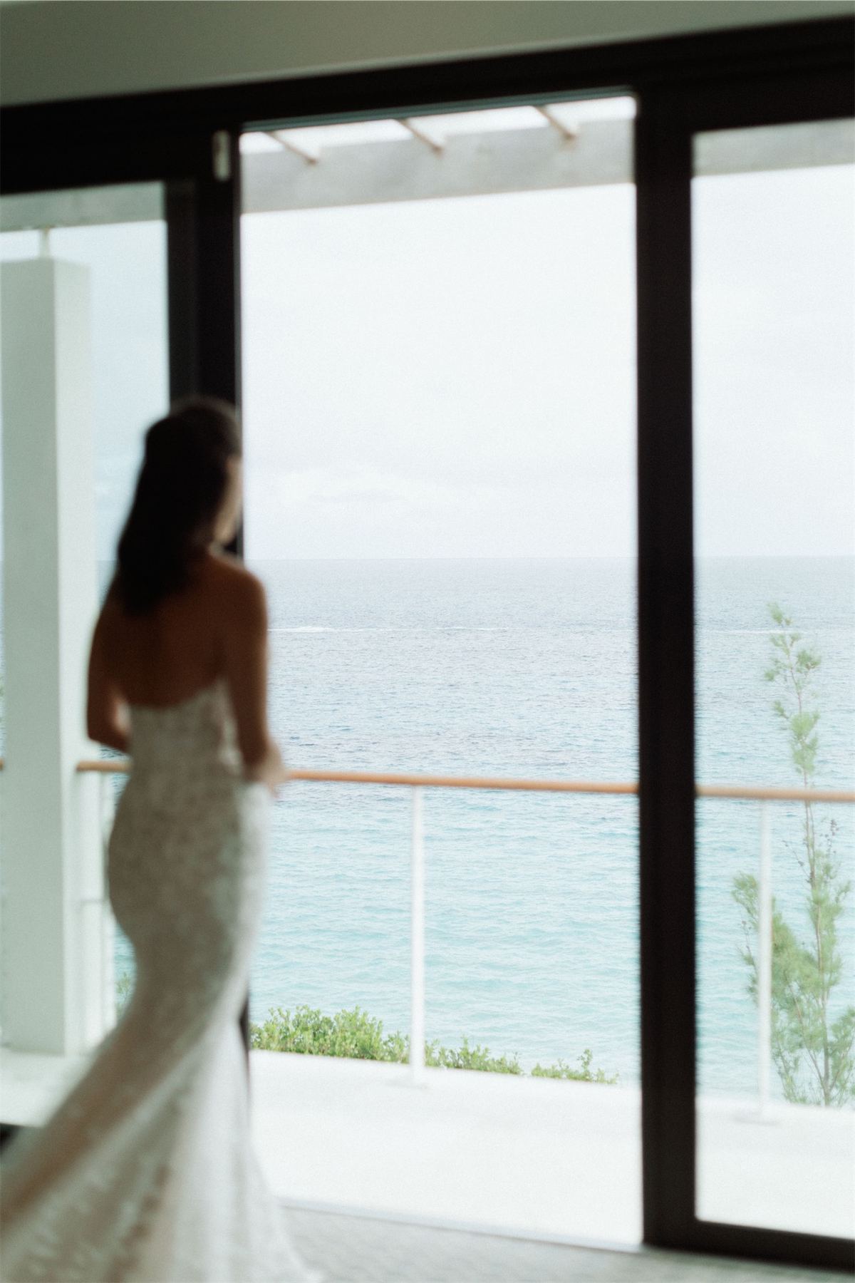 Bride in a fitted lace gown looking out an open window toward the ocean