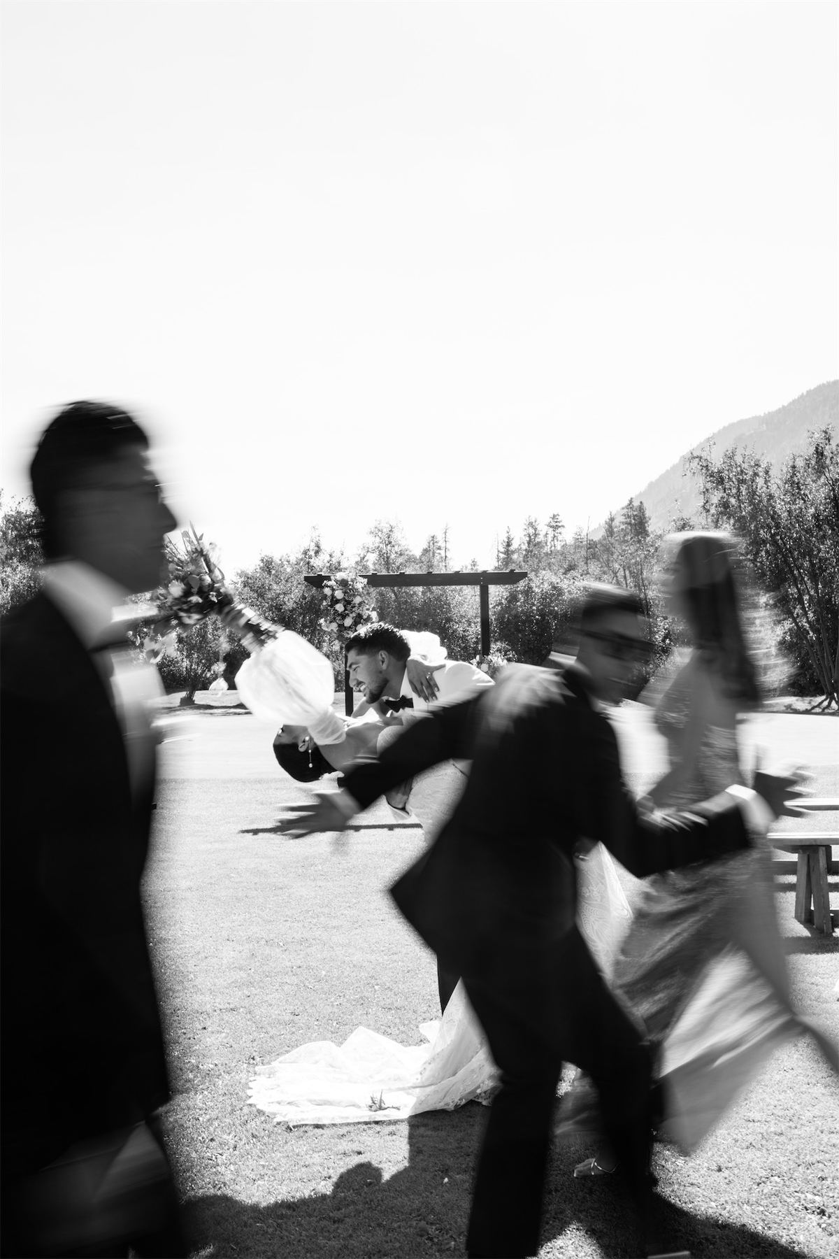 Black and white motion-blurred frame of the couple and guests crossing a sunlit drive
