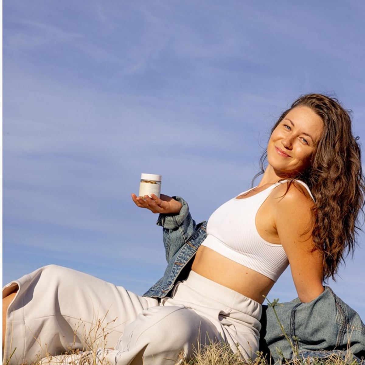 Model in cream separates against blue sky, a capsule jar balanced in her palm