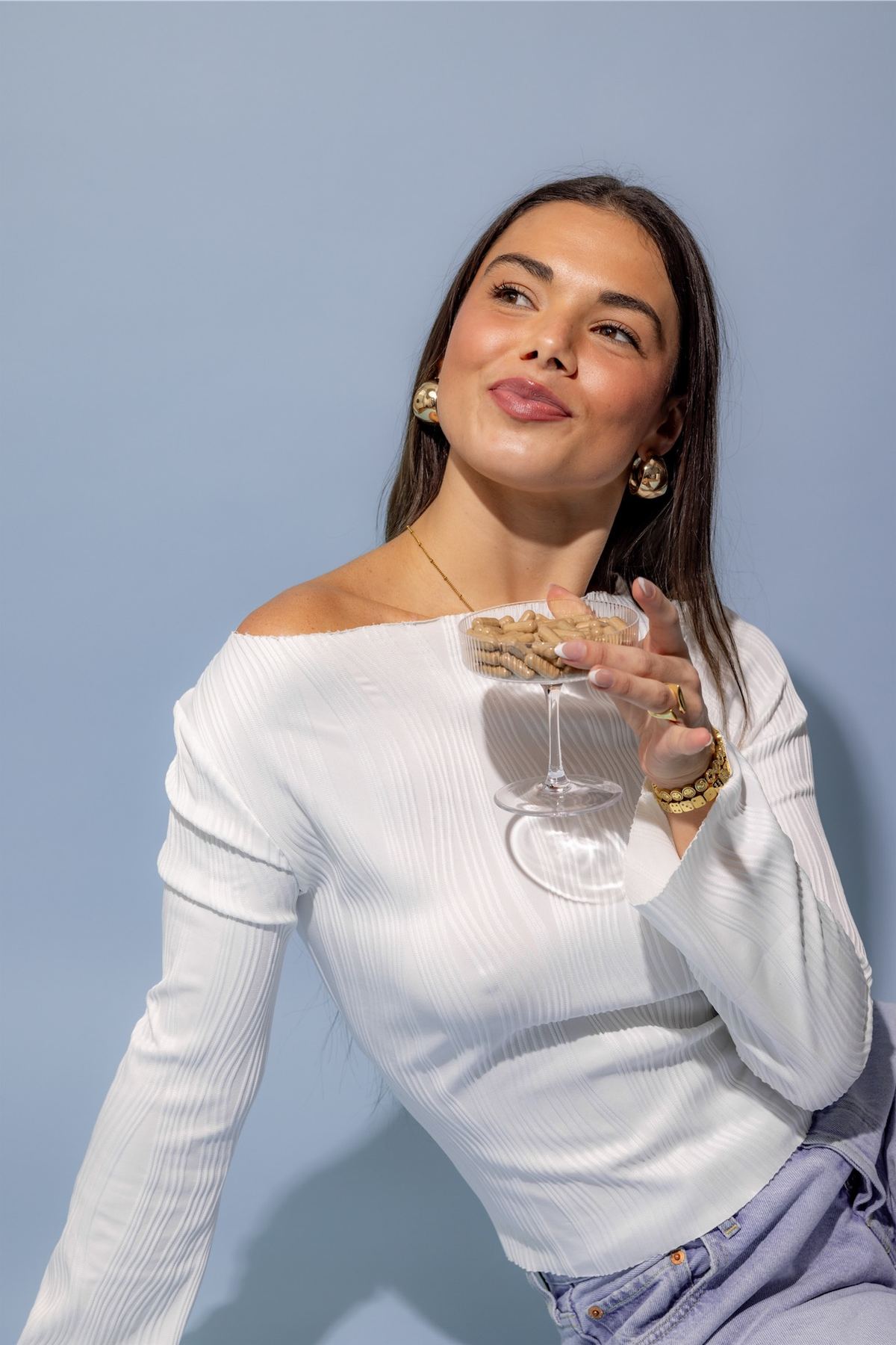 Model in a white knit top smiling with a glass and capsule jar at a lilac wall
